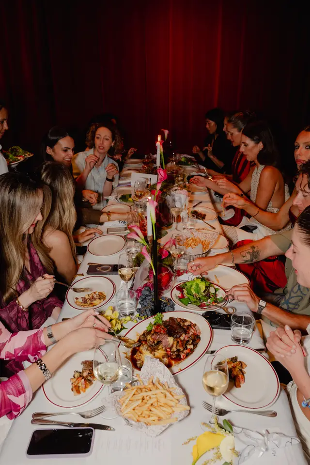 An overhead shot captures multiple people, mostly women, dining at a long table in a dimly lit room. The table is laden with various dishes, wine glasses, candles, and floral arrangements, suggesting a lively and elegant social event. Red curtains serve as a backdrop, adding to the intimate atmosphere.