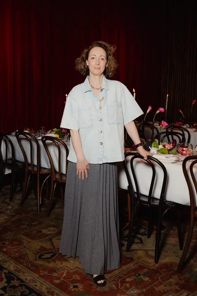 A full-length portrait of a woman standing in a formal event setting. She is wearing a light blue, short-sleeved button-up shirt and a long grey pleated skirt, leaning casually on a dark wooden chair. Behind her, a long table is set with white tablecloths, floral arrangements featuring pink calla lilies, pomegranates, grapes, and lit candles, all against a backdrop of dark red curtains and a patterned rug.
