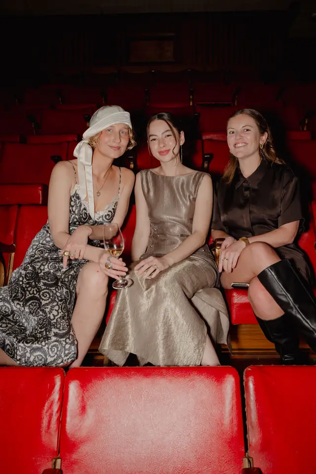 Three women are seated in a row of red theater seats, looking towards the camera and smiling. They are dressed in elegant attire, suggesting attendance at a special event. The background shows more rows of empty red seats, indicating a theater or auditorium setting.