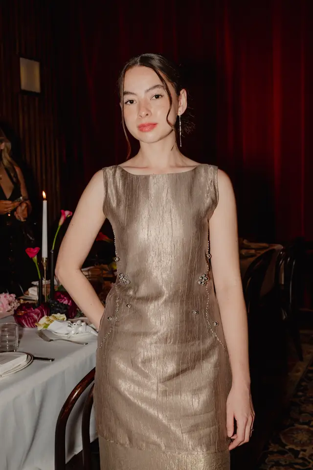 A young woman in a shimmering gold dress with silver embellishments stands in a dimly lit room, looking directly at the camera. Behind her, a set table with lit candles and floral arrangements suggests an elegant event. Dark red curtains form the backdrop, contributing to a warm and sophisticated atmosphere.