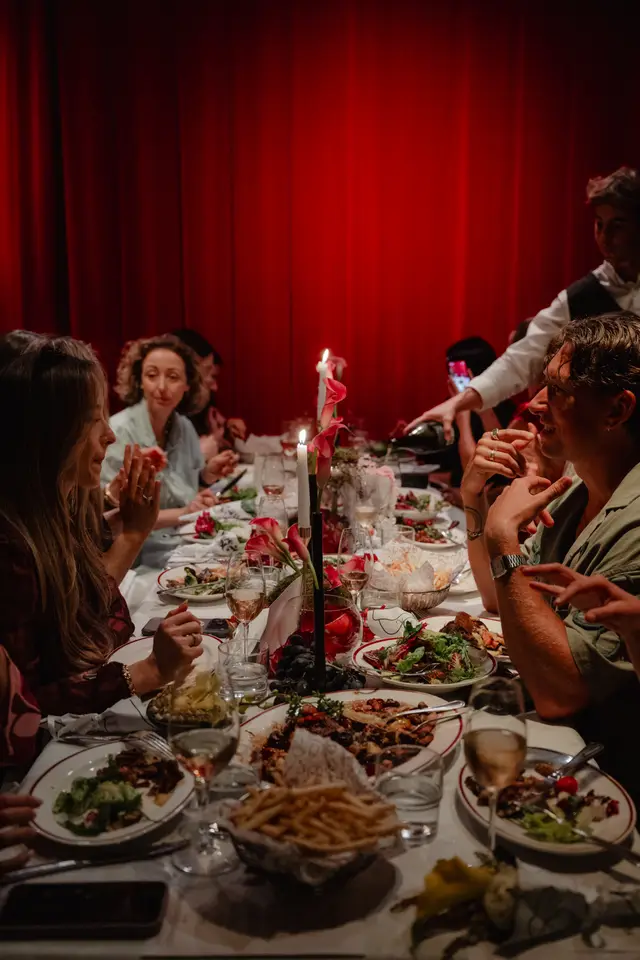 An intimate, low-light photograph captures a lively dinner party, with multiple guests seated around a long table. The table is adorned with various dishes, wine glasses, lit candles, and elegant floral arrangements, set against a dramatic red curtain backdrop. A server is visible pouring wine, while guests engage in conversation and enjoy their meal.