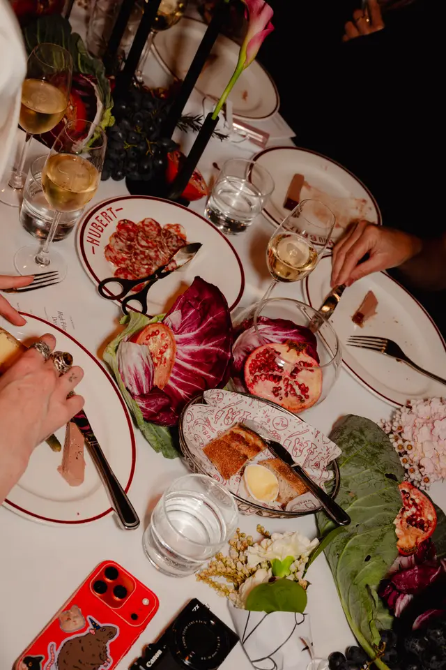 A top-down view captures an elegant dinner table setting, featuring various dishes, drinks, and decorative elements. Multiple hands are visible, interacting with the food and cutlery, suggesting a lively social gathering. The table is adorned with fresh produce, flowers, and fine dining accoutrements.