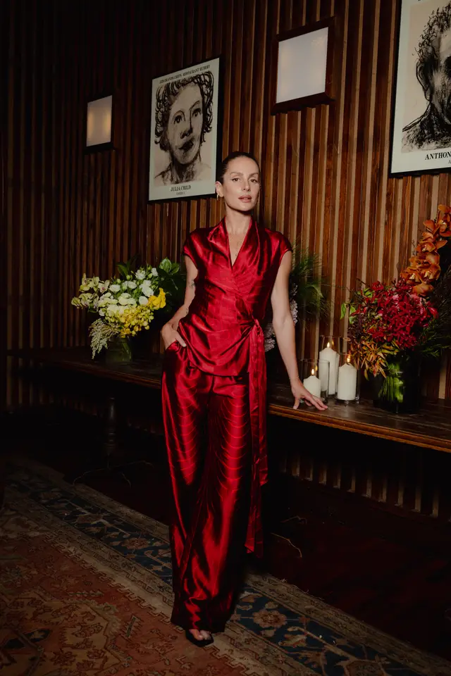 A woman in a vibrant red satin wrap top and matching wide-leg pants stands confidently in a dimly lit room. She is positioned between two large floral arrangements and several lit candles on a dark wooden surface, against a slatted wooden wall adorned with framed artwork.