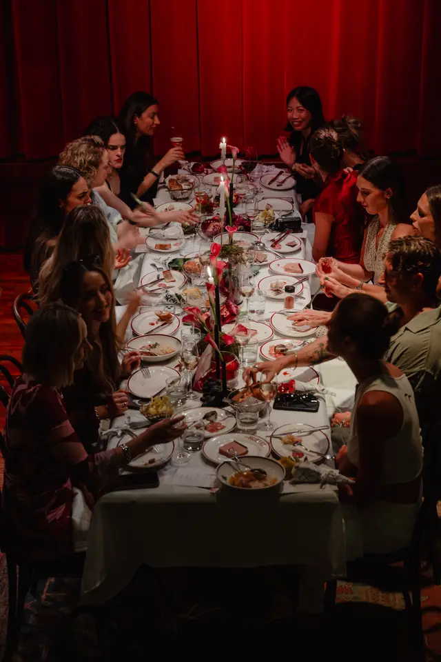 A high-angle shot captures a group of people, predominantly women, gathered around a long, candlelit dining table. The table is laden with food, drinks, and floral arrangements, suggesting an intimate and festive dinner event. Red curtains form the backdrop, adding to the warm and sophisticated atmosphere.