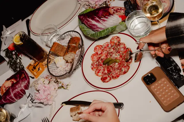 An overhead shot captures a vibrant dining table spread with various dishes and drinks, suggesting a social event or upscale meal. Two sets of hands are visible, interacting with the food, including a plate of sliced salami with pickles and a basket of bread. The table is elegantly set with white linen, fresh flowers, and other culinary elements.