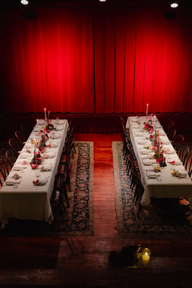 An overhead shot captures two long, elegantly set dining tables arranged in front of a dramatic red velvet stage curtain. Each table is adorned with elaborate floral centerpieces, an array of lit candles, individual place settings, and decorative fruit. The dark wooden floor, partially covered by patterned rugs, adds to the sophisticated and intimate ambiance of the event space.
