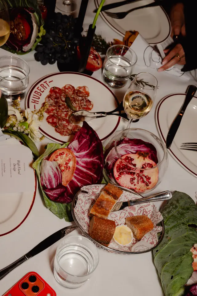 A top-down view of an elegantly set dining table featuring various dishes, drinks, and floral arrangements. Plates of charcuterie, bread with butter, and fresh produce like pomegranates, radicchio, and grapes are visible, alongside wine and water glasses. A menu card for a 'High Floral Design' event at 'Restaurant Hubert' is also present.