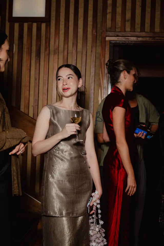 A young woman in a metallic gold dress holds a wine glass and a decorative bag, looking to her left with a slight smile. She is positioned in the foreground, with other guests partially visible in the background against a warm, wood-paneled wall. The scene suggests a social gathering or event.