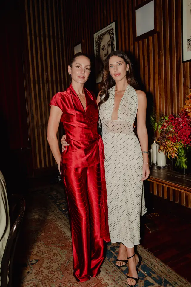 Two women pose for a full-length photo at an indoor event. The woman on the left wears a red satin wrap top and matching wide-leg pants, while the woman on the right wears a cream-colored, polka-dotted halter dress with a deep V-neck. They stand in front of dark wood paneling, with a floral arrangement and candles visible on a table to the right.
