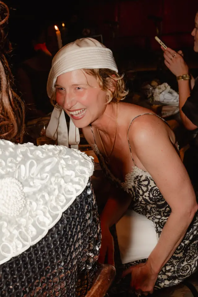 A candid, close-up shot of a woman with short blonde hair and a cream-colored hat, laughing heartily while looking towards another person. She is wearing a patterned dress and a delicate gold necklace, captured in a dimly lit indoor setting with hints of other attendees and candlelight in the background.