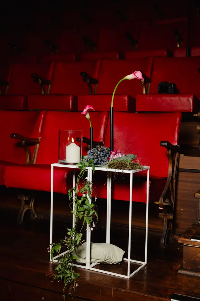 A sophisticated floral and candle arrangement is displayed on a white metal side table, set against a backdrop of empty, plush red theater seats. Two elegant pink and white calla lilies stand in slender black vases, accompanied by dark grapes, moss, and a lit white candle in a glass hurricane. A green vine with small pink flowers trails gracefully down the side of the table, adding a natural touch to the dramatic setting.