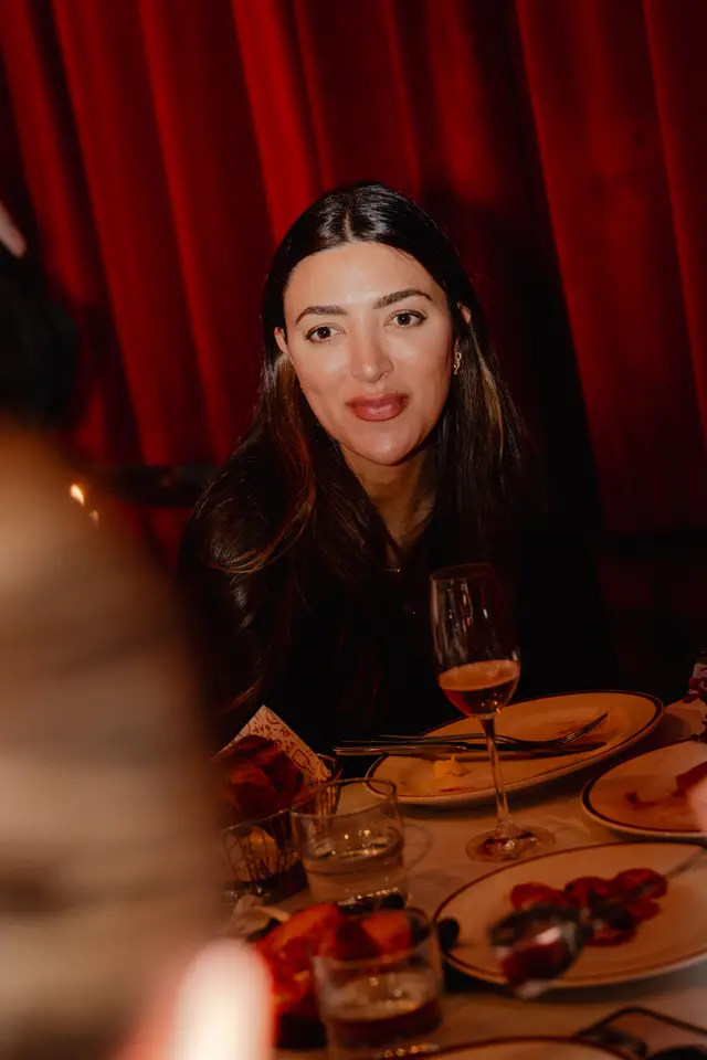 A woman with long dark hair is seated at a dinner table, looking directly at the camera with a subtle smile. The background features rich, draped red curtains, and the table is set with plates, cutlery, and glasses, indicating a social gathering or meal. The lighting is warm and creates an intimate atmosphere.