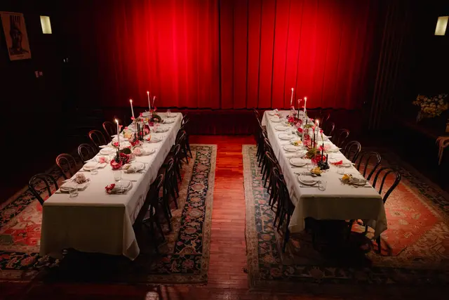 Two long, elegantly set dinner tables with white tablecloths, lit candles, and vibrant floral arrangements are positioned in a dimly lit room. A dramatic red curtain serves as the backdrop, creating an intimate and luxurious atmosphere. The tables are flanked by dark wooden chairs and rest on patterned rugs over a wooden floor.