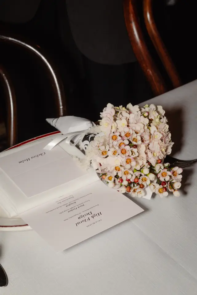 A close-up, overhead shot of an elegant table setting at an event, featuring a small, delicate bouquet of light pink and white flowers. A white menu card, titled 'High Floral Design' and 'Fig & Bloom', and a place card for 'Chelsea Holm' are neatly arranged on a white tablecloth, with a dark, out-of-focus background showing parts of a wooden chair.