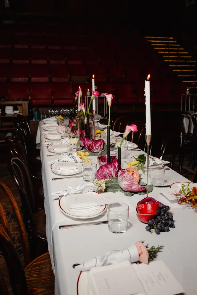 A long event table is elegantly set with white tablecloths, individual place settings, and a vibrant centerpiece arrangement. The centerpiece features pink calla lilies, radicchio, pomegranates, and grapes, interspersed with tall white candles in dark candlesticks. In the background, rows of dark red theater-style seating are visible, suggesting an event in a unique venue.