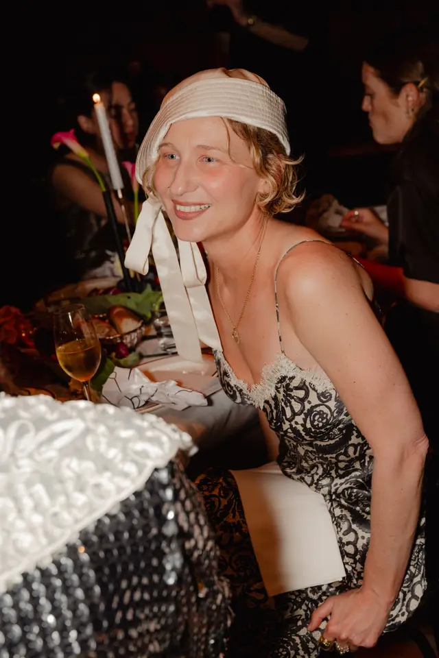 A smiling woman with blonde, curly hair and a cream-colored hat is captured mid-conversation at a dimly lit event. She is wearing a patterned slip dress with lace trim and a gold necklace, with a wine glass and a table setting visible in the foreground. Other guests are blurred in the background, contributing to an intimate and social atmosphere.