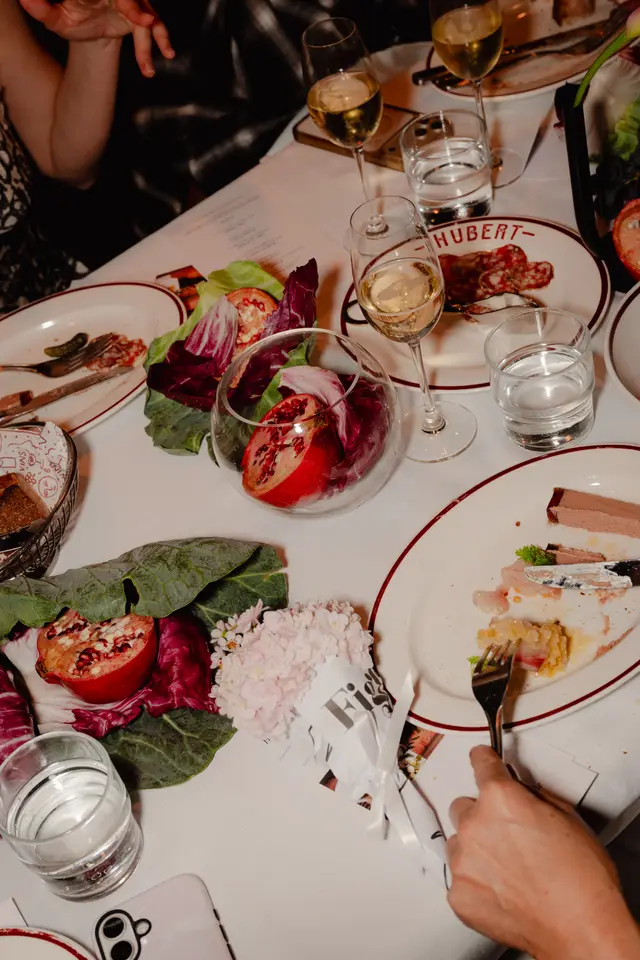 A top-down view of an elegant dining table set with a white tablecloth, featuring partially eaten plates of food, wine, and water glasses. Decorative elements include halved pomegranates, various lettuce leaves, and a small bouquet of light pink flowers. Multiple hands are visible, indicating a social dining experience.