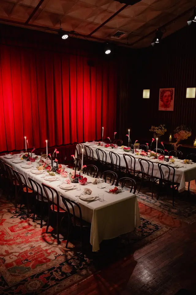 The image captures an elegantly set event space featuring two long dining tables draped in white tablecloths, adorned with floral arrangements, candles, and fruit. A vibrant red curtain serves as a dramatic backdrop, illuminated by spotlights, while patterned rugs and dark wooden floors complete the sophisticated ambiance. The tables are fully set with plates, glasses, and napkins, ready for guests.