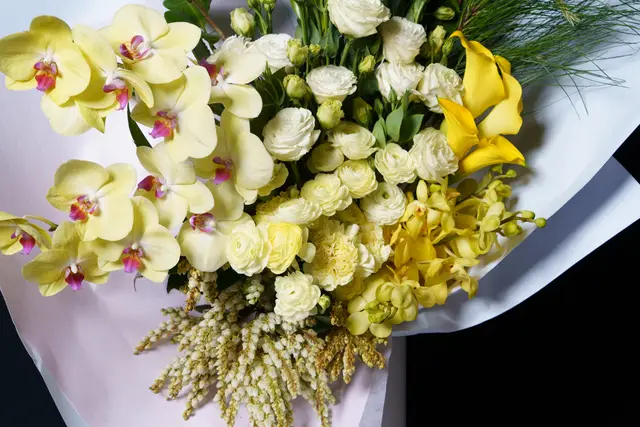A vibrant, close-up shot of a large floral bouquet featuring a variety of yellow and white flowers with green foliage, wrapped in white paper. The arrangement includes prominent yellow orchids, white ranunculus, and yellow calla lilies, set against a dark background. The composition highlights the textures and colors of the diverse blooms.