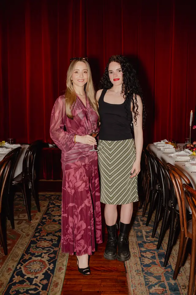 Two women are standing side-by-side, smiling at the camera, in an indoor event setting. The woman on the left wears a long, sheer, floral-patterned dress in shades of pink and purple, holding a wine glass. The woman on the right wears a black tank top, a green and white striped skirt, and black boots. Behind them are rich red curtains and to their sides are set dining tables with chairs, suggesting a formal gathering.