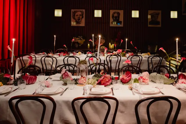 A long dining table is elegantly set for an event, featuring white tablecloths, dark bentwood chairs, and multiple place settings. The table is adorned with a rich centerpiece of pink calla lilies, radicchio, grapes, and halved pomegranates, interspersed with lit taper candles in dark holders. The background is dimly lit, showing dark walls, red curtains, and framed portraits, creating an intimate and sophisticated ambiance.