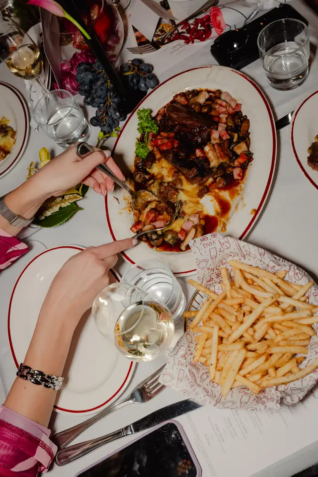An overhead shot captures a lively dinner table scene where two sets of hands are serving a rich main course of beef, mushrooms, and bacon from a large platter onto a plate. The table is elegantly set with a white tablecloth, various drinks, a basket of french fries, and floral decorations, suggesting a social dining event.