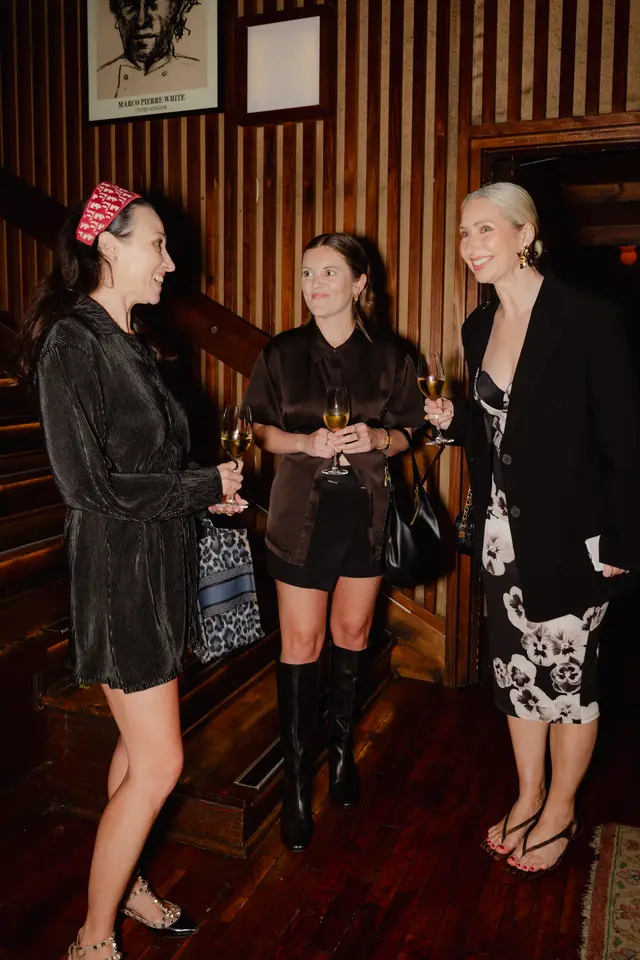 Three women are captured mid-conversation at an indoor event, each holding a glass of champagne. They are dressed in elegant evening attire, standing in a dimly lit space with rich, dark wooden paneling and a visible staircase in the background. A framed portrait of Marco Pierre White hangs on the wall.