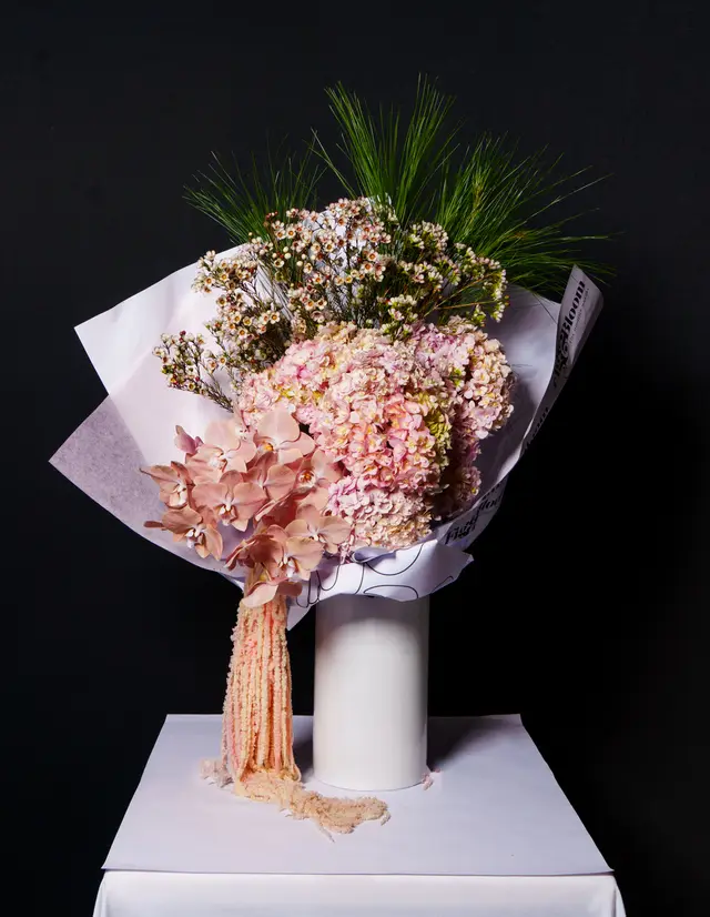 A large, elaborate floral bouquet featuring pink hydrangeas, peach orchids, small white and pink filler flowers, and long green pine needles. The bouquet is wrapped in white and translucent paper, resting in a tall white cylindrical vase on a white draped surface against a dark background. A long, textured pink-orange floral element drapes from the bouquet onto the table.