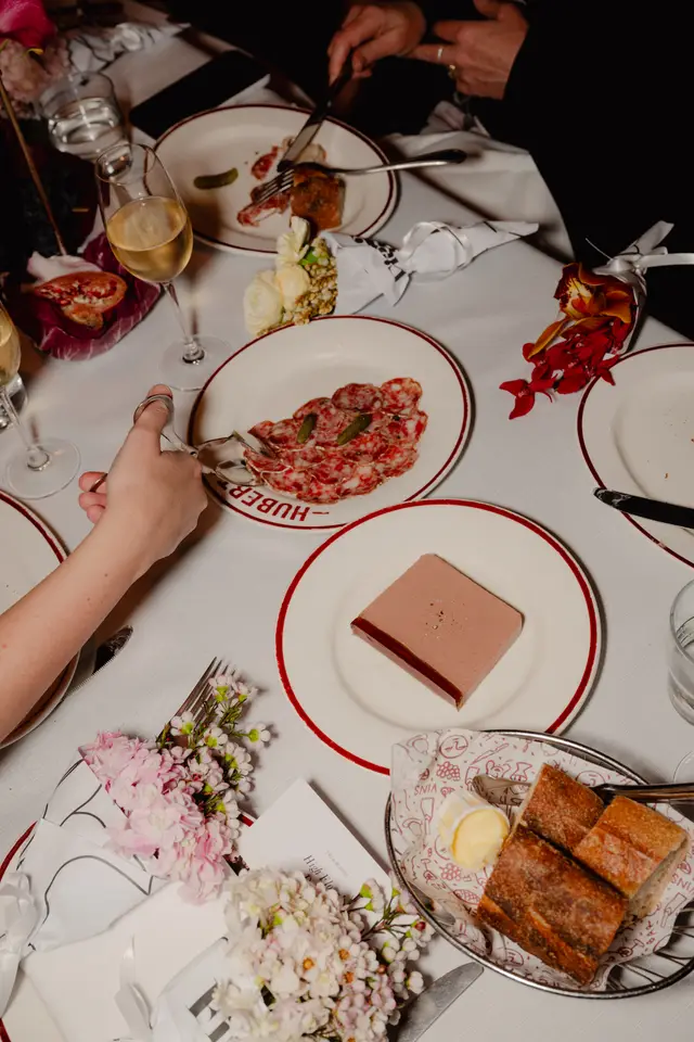 An overhead view of a sophisticated dining table set for an event, showcasing various gourmet dishes, wine glasses, and elegant floral arrangements. Multiple hands are visible, interacting with the food and table setting, suggesting a lively and intimate gathering. The scene is well-lit, highlighting the details of the food and decor.