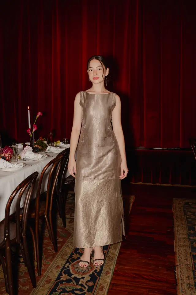 A woman in a shimmery gold, sleeveless dress stands facing the camera, smiling slightly, against a backdrop of rich red velvet curtains. To her left, a formal dining table is set with a white tablecloth, dark wooden chairs, a lit white candle, and elegant floral arrangements featuring pink calla lilies and other blooms. The scene depicts an upscale evening event.