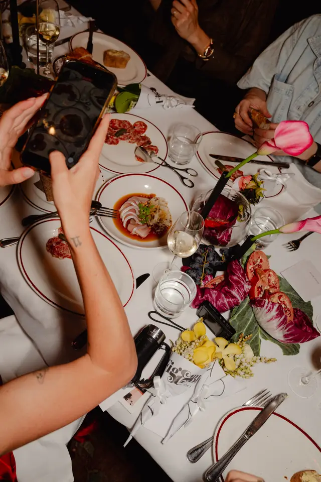 An overhead shot captures a lively dinner table scene, featuring various gourmet dishes, drinks, and decorative floral arrangements. A person's hands are prominently shown taking a photo of the table with a smartphone, while other guests' hands are visible interacting with food and drinks. The setting suggests an elegant social event.