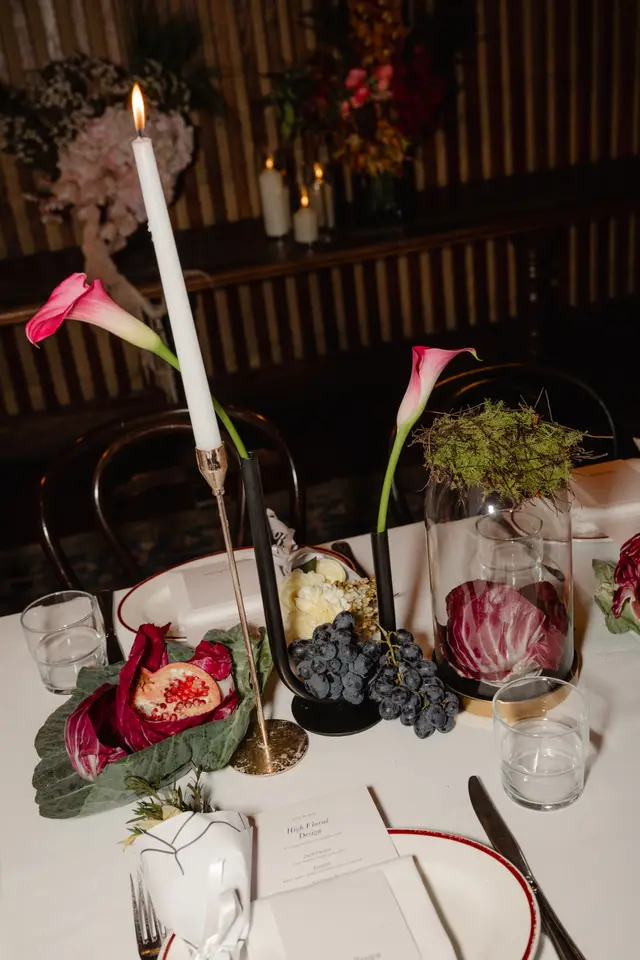 A close-up shot of an elegantly set table at an evening event, featuring a lit white taper candle in a gold holder, two pink calla lilies, and various fruits and vegetables as part of the centerpiece. Place settings with white plates, red-rimmed saucers, menu cards, glassware, and cutlery are also visible, all set on a white tablecloth against a dark, warm-toned background.