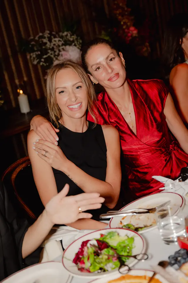 Two smiling women are captured in a close-up shot at an indoor event, seated at a dining table with food and drinks. The woman on the left, in a black top, has her arm around the woman on the right, who wears a red patterned dress and gold jewelry. Blurred floral arrangements and a lit candle are visible in the background, suggesting an elegant, social gathering.