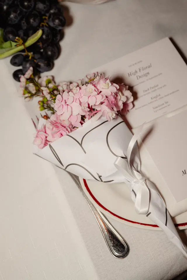 A close-up, overhead shot of a formal table setting featuring a small bouquet of pink and white flowers wrapped in white paper with a dark pattern and tied with a white ribbon. Beside the bouquet, a menu card titled "High Floral Design" is partially visible, along with a white plate with a red rim and a silver fork. A bunch of dark grapes rests in the upper left corner, all set on a white tablecloth.