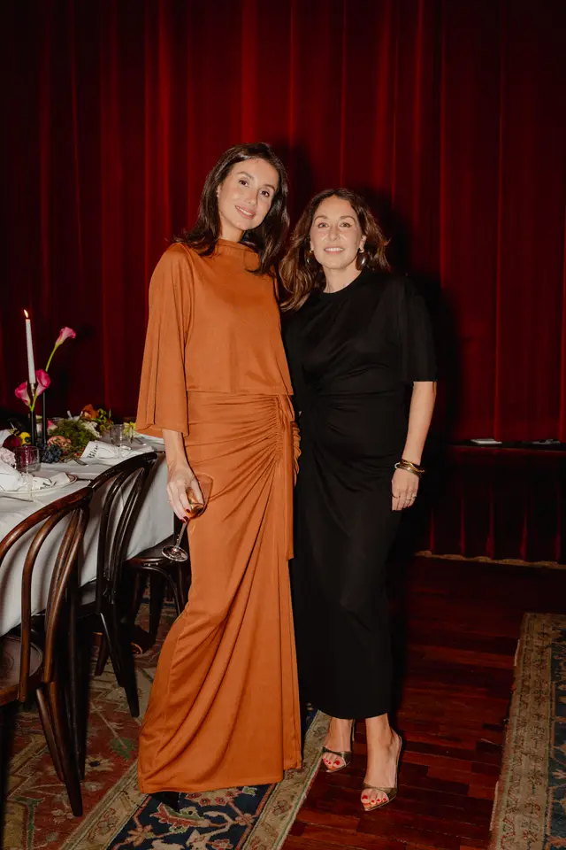 Two women are posing for a full-length photo at an indoor event. The woman on the left wears a long-sleeved rust-colored dress and holds a wine glass, while the woman on the right wears a black dress. In the background, a set table with floral arrangements and a lit candle is visible, against rich red velvet curtains.