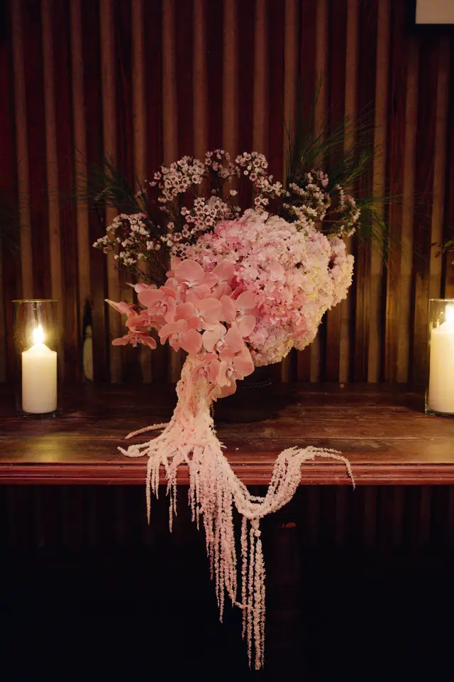 A luxurious floral arrangement featuring a prominent display of pink orchids, light pink hydrangeas, and cascading pink amaranthus. The arrangement is centered on a dark wooden surface, flanked by two lit pillar candles in clear glass holders. The background consists of dark, vertically paneled walls, creating an intimate and elegant atmosphere.