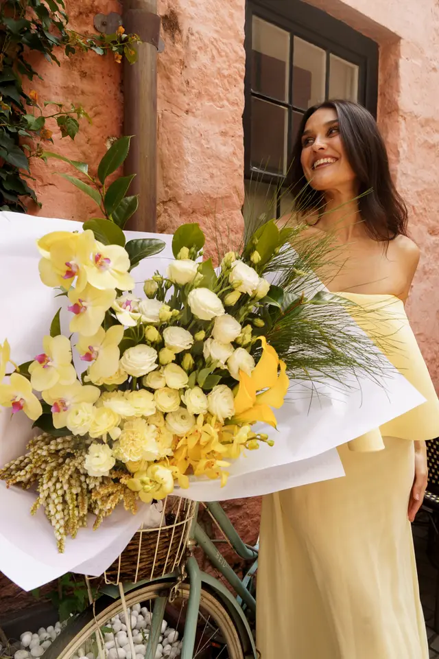 A smiling woman in a yellow off-the-shoulder dress stands next to a vintage-style bicycle, holding a large, vibrant bouquet of yellow and white flowers. The scene is set outdoors against a textured peach-colored building wall with a dark window and some green foliage. The bouquet is prominently featured, wrapped in white paper and resting in the bicycle's front basket.