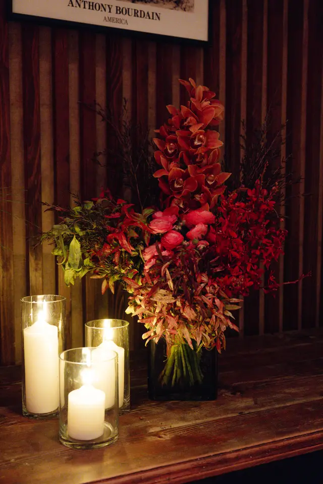 A large, vibrant floral arrangement featuring red and orange orchids, other red flowers, and various green and reddish foliage is prominently displayed in a dark vase. Three lit white pillar candles in clear glass holders sit on a dark wooden surface in the foreground, creating a warm, intimate atmosphere. A dark, vertically paneled wooden wall serves as the background, with a framed picture partially visible at the top.
