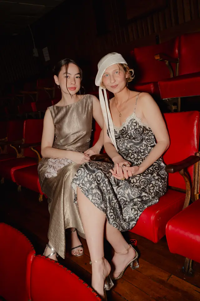 Two women are seated closely together in a row of red theater seats. The woman on the left wears a shimmering gold dress, while the woman on the right wears a patterned black and white slip dress with a white lace trim and a distinctive white hat. Their hands are clasped together, and they both look towards the viewer with a gentle expression, illuminated by soft lighting against a dark background.