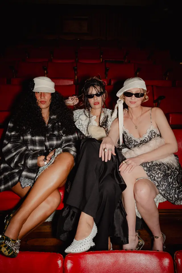 Three women are seated in a row of red theater seats, looking towards the camera. They are dressed in stylish, eclectic outfits, including a plaid shirt, a lace top, and a patterned dress, accessorized with hats, sunglasses, and prominent jewelry. The setting is dimly lit, highlighting the vibrant red of the seats and the details of their attire.