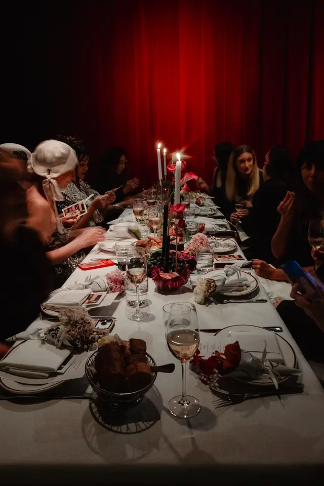 A long dining table is set for an event, illuminated by candlelight against a rich red curtain backdrop. Multiple guests are seated, engaging with the table settings and each other. The table is adorned with floral arrangements, candles, and place settings, creating a warm and intimate atmosphere.