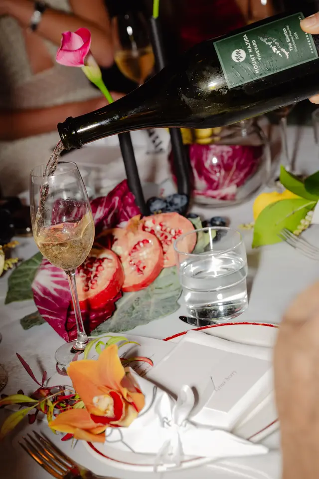 A close-up shot captures white wine being poured from a dark bottle into a stemmed glass, creating a dynamic splash. The elegant table setting is adorned with vibrant elements including sliced pomegranates, red radicchio leaves, dark grapes, and a bright yellow lemon. Floral accents, such as an orange orchid and a pink calla lily, enhance the luxurious atmosphere.