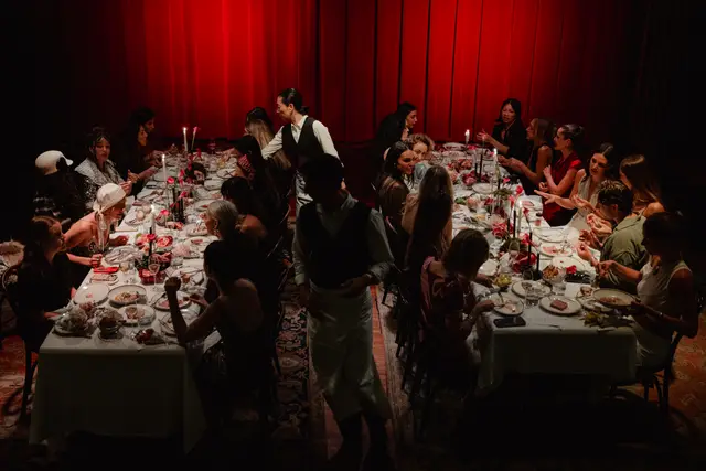 An overhead shot captures a long, elegantly set dinner table filled with multiple guests and staff, illuminated by candlelight. The scene is set against a dramatic backdrop of deep red curtains, creating an intimate and sophisticated atmosphere. Various dishes, wine glasses, and floral arrangements adorn the white tablecloth.