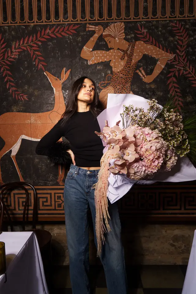 A woman stands holding a large, elaborate bouquet of pink and peach flowers against a dark wall adorned with ancient Greek-style murals. She is wearing a black ribbed top with feathered cuffs and blue jeans, looking slightly upwards and to the right. The bouquet features hydrangeas, orchids, and cascading amaranthus, wrapped in white and light purple paper.