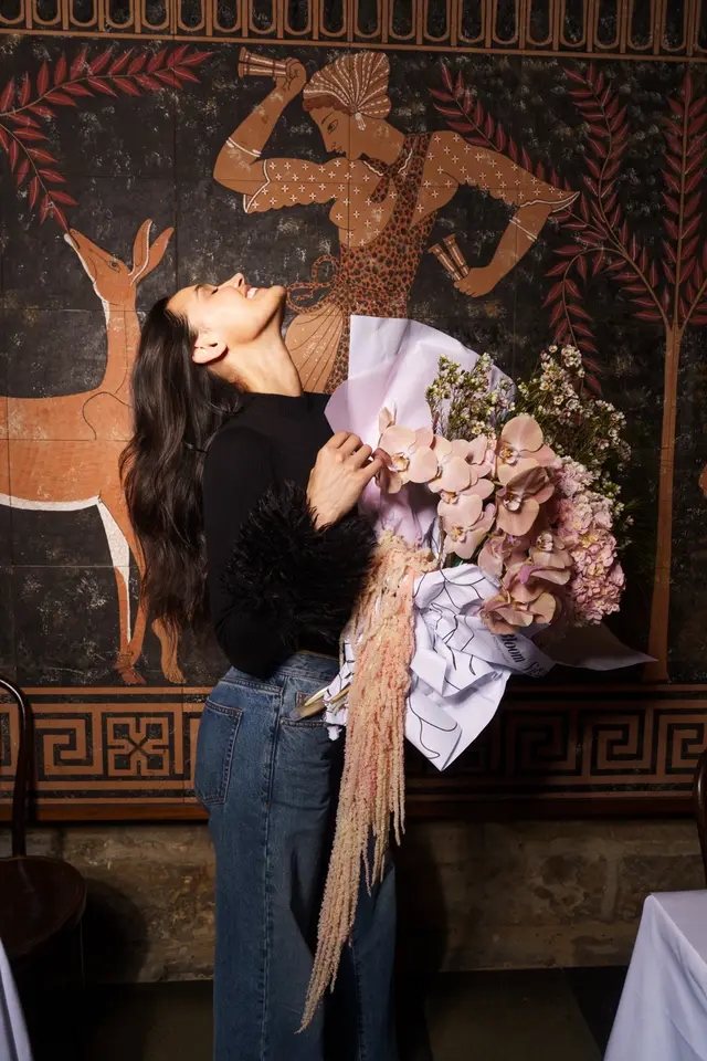 A woman with long dark hair, wearing a black top with feathered cuffs and blue jeans, smiles joyfully while holding a large, artfully arranged bouquet of flowers. She is positioned against a dark wall featuring a stylized mural with a deer and a human figure. The bouquet includes pink orchids, pink hydrangeas, and long, feathery pink amaranth, wrapped in pink and white paper.