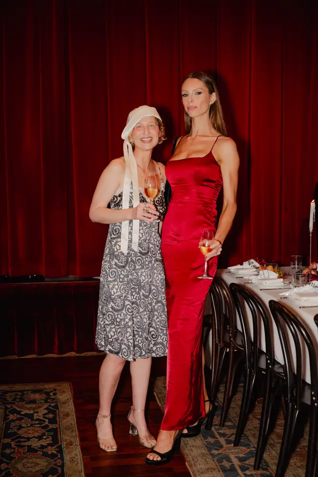 Two women are captured full-length at an indoor event, holding glasses of white wine. They stand in front of a deep red curtain backdrop, with a formally set dining table and chairs visible to their right. The woman on the left wears a patterned dress and a light-colored head covering, while the woman on the right wears a long, red satin slip dress.