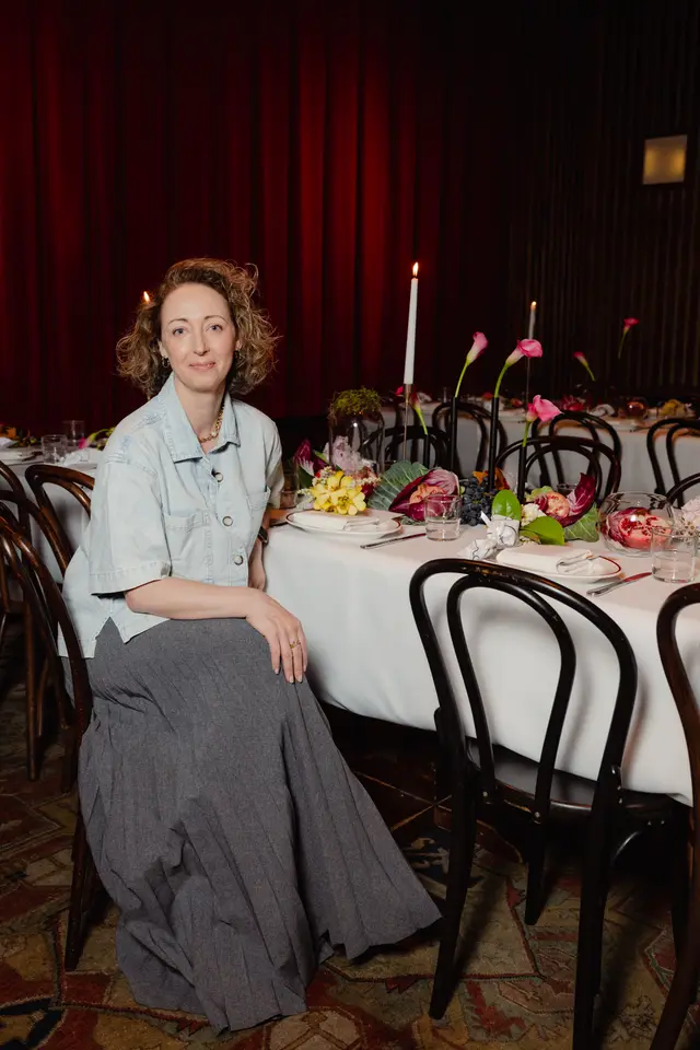 A woman with curly blonde hair and a light blue denim-style shirt sits at a beautifully decorated dining table, looking towards the camera. The table is adorned with floral arrangements, candles, fruit, and place settings, set against a backdrop of dark red curtains. The scene suggests an elegant launch event or dinner.