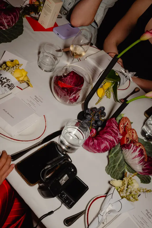 A top-down view of a beautifully set dining table at an event, featuring multiple place settings, various food items like radicchio, grapes, and pomegranate, and elegant floral arrangements. Several hands and arms are visible, suggesting guests are seated and interacting with the table. Menus for a 'High Floral Design' event are also present.