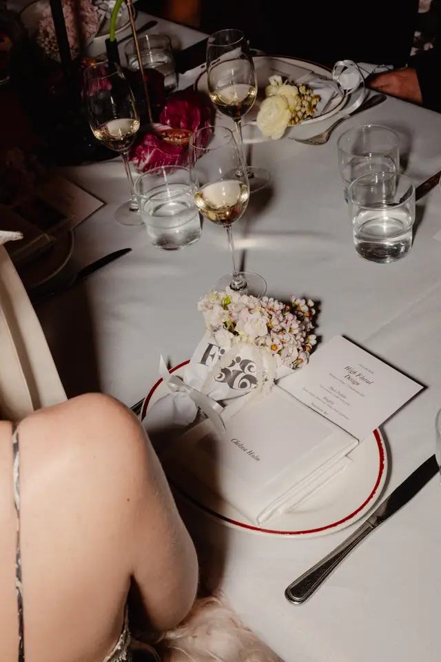 A close-up, overhead shot of a formal dining table setting at an event. The table is adorned with white linen, wine glasses, water glasses, cutlery, and floral arrangements. A menu for 'Fig & Bloom High Floral Design' is prominently displayed, along with a small wrapped gift or favor.