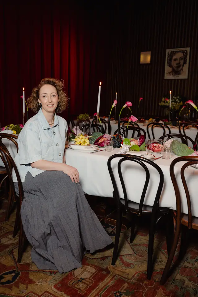 A woman with curly hair, wearing a light denim shirt and a grey skirt, sits smiling at a long, elegantly set dinner table. The table is adorned with a white tablecloth, candles, various flowers, fruits, and vegetables, suggesting a sophisticated event. In the background, rich red curtains and dark wood paneling create a warm, intimate atmosphere.